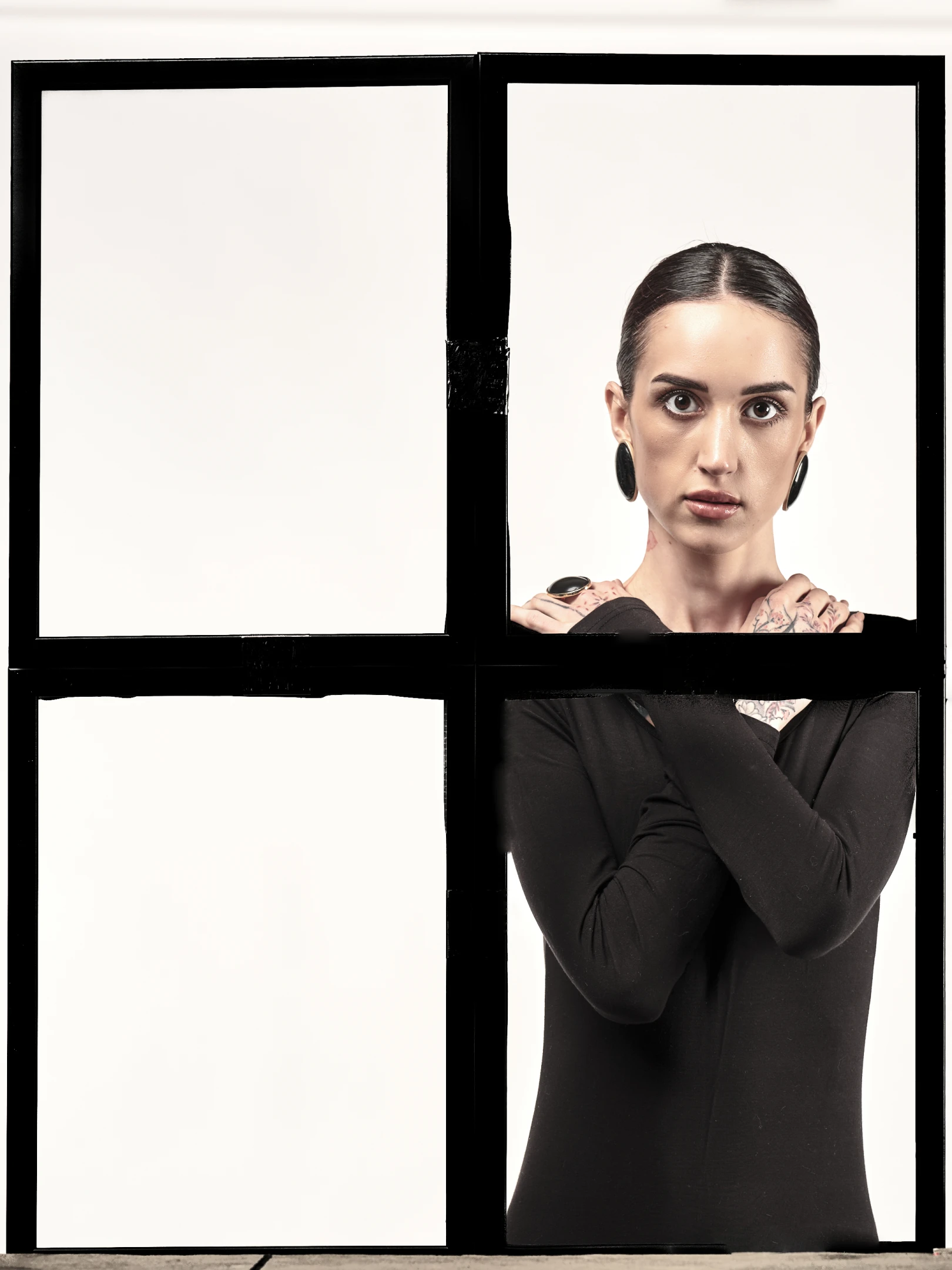 The portrait features a woman with dark, sleekly pulled-back hair, bold dark eyes, and a neutral, direct gaze that engages the viewer with an intense, almost confrontational stare. Her expression is stoic, with lips slightly parted and a subtle tension in her jawline, suggesting resolve or defiance. She wears a form-fitting black long-sleeve top, accentuating her arms and shoulders, which are adorned with intricate tattoos—visible on her neck, arms, and hands, including a large ring on her right hand and thick, dark hoop earrings. Her arms are crossed over her chest, a posture that conveys confidence or guardedness. The lighting is high-contrast, spotlighting her face and upper body again...