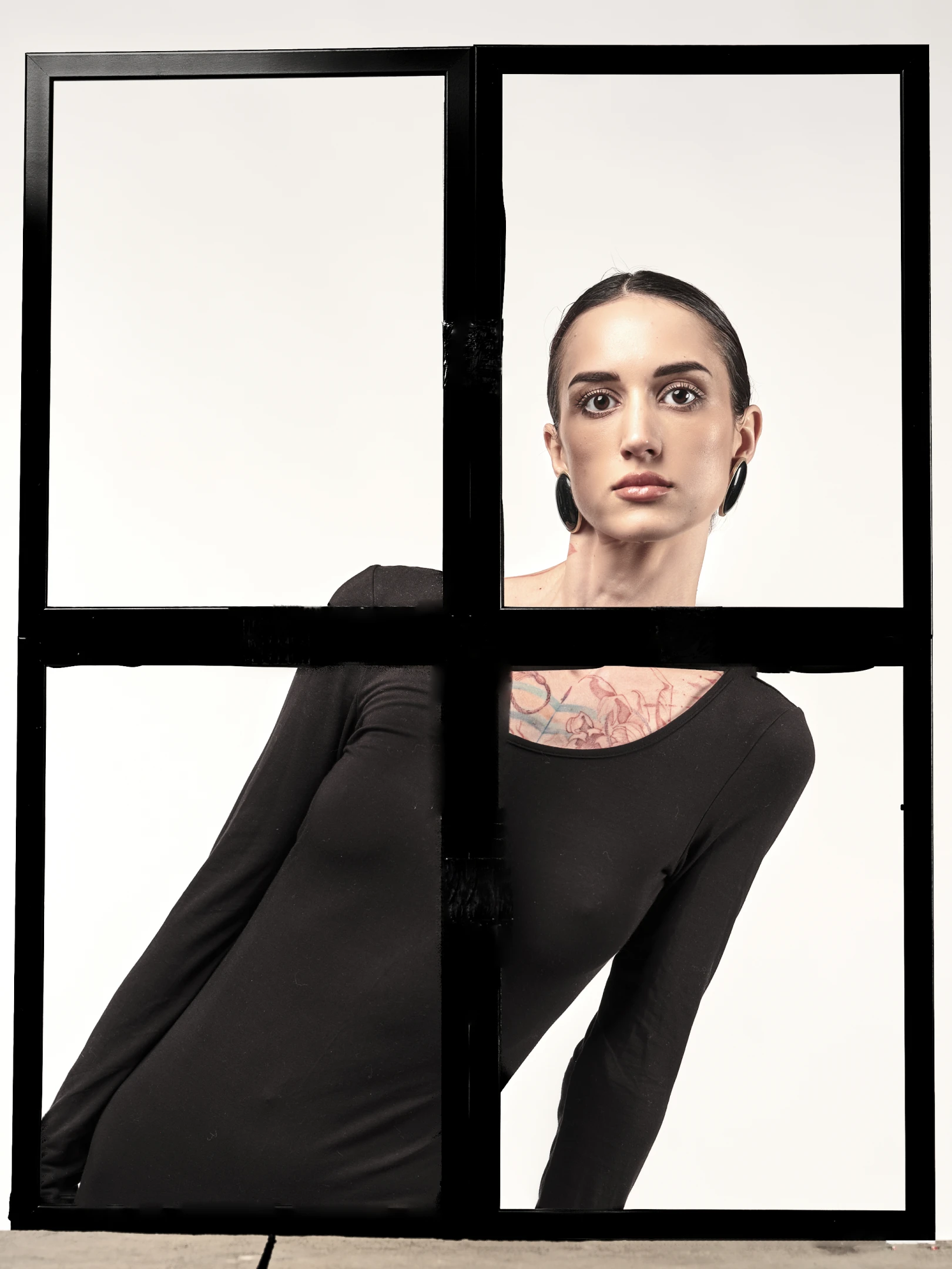 The portrait features a woman with dark, sleek hair pulled back, revealing a neutral, direct gaze that conveys confidence and calmness. Her face is framed by a minimalist black windowpane structure, divided into four quadrants, with her head and shoulders visible in the top-right panel and her torso and arms occupying the bottom-left and bottom-right panels. She wears a form-fitting black long-sleeve top, accentuating her silhouette, and large, dark hoop earrings that add a bold, modern touch. A visible tattoo adorns her neck and upper chest, featuring floral or abstract patterns in muted tones, contrasting with the stark black of her attire. The lighting is soft and even, highlighting he...