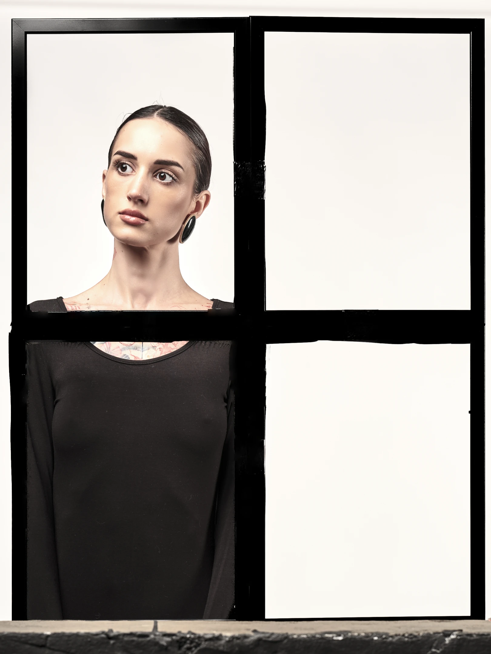 The portrait captures a woman with dark, sleekly pulled-back hair, wearing large, dark hoop earrings that contrast with her fair skin. Her expression is neutral yet contemplative, with her gaze directed slightly upward and to the left, suggesting introspection or observation. She is dressed in a simple black long-sleeved top, revealing a subtle floral tattoo just beneath the neckline, adding an element of personal artistry. The lighting is soft and even, highlighting her facial features with a clinical clarity, while casting gentle shadows that emphasize her jawline and neck. The background is a stark, minimalist white, framed by a bold black grid that divides the image into four quadrant...