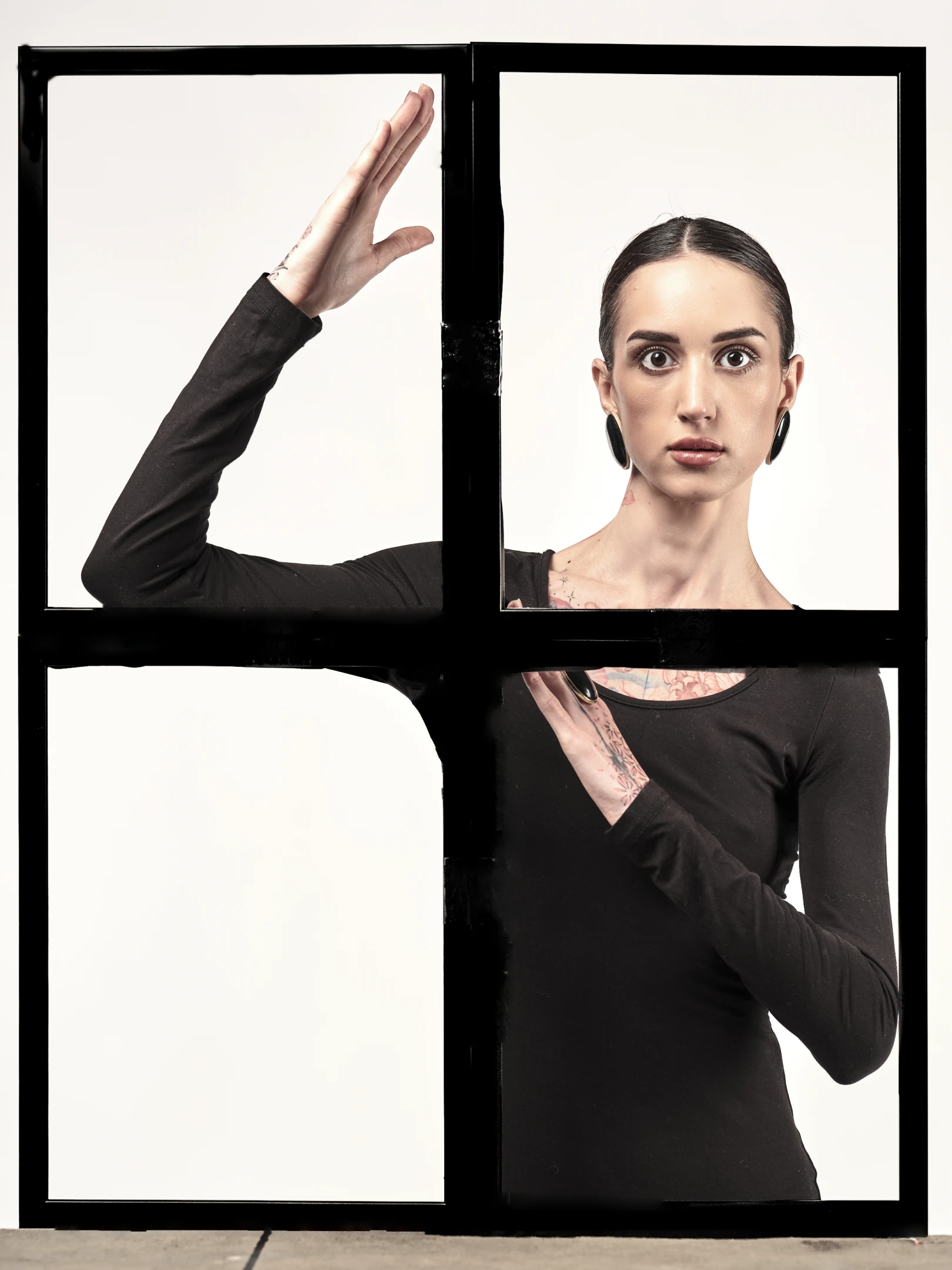 The portrait features a young woman with dark, sleek hair pulled back from her face, revealing bold, expressive eyes and a neutral, direct gaze that engages the viewer. Her skin tone is fair, and she wears large, dark hoop earrings that add a bold contrast to her otherwise minimalist look. She is dressed in a form-fitting black long-sleeve top, which accentuates her tattoos visible on her neck, shoulders, and arms — intricate designs that hint at personal expression and artistry. Her pose is dynamic yet composed: one arm reaches upward with fingers slightly spread, while the other rests near her chest, suggesting movement frozen in time. The lighting is soft and even, illuminating her fea...