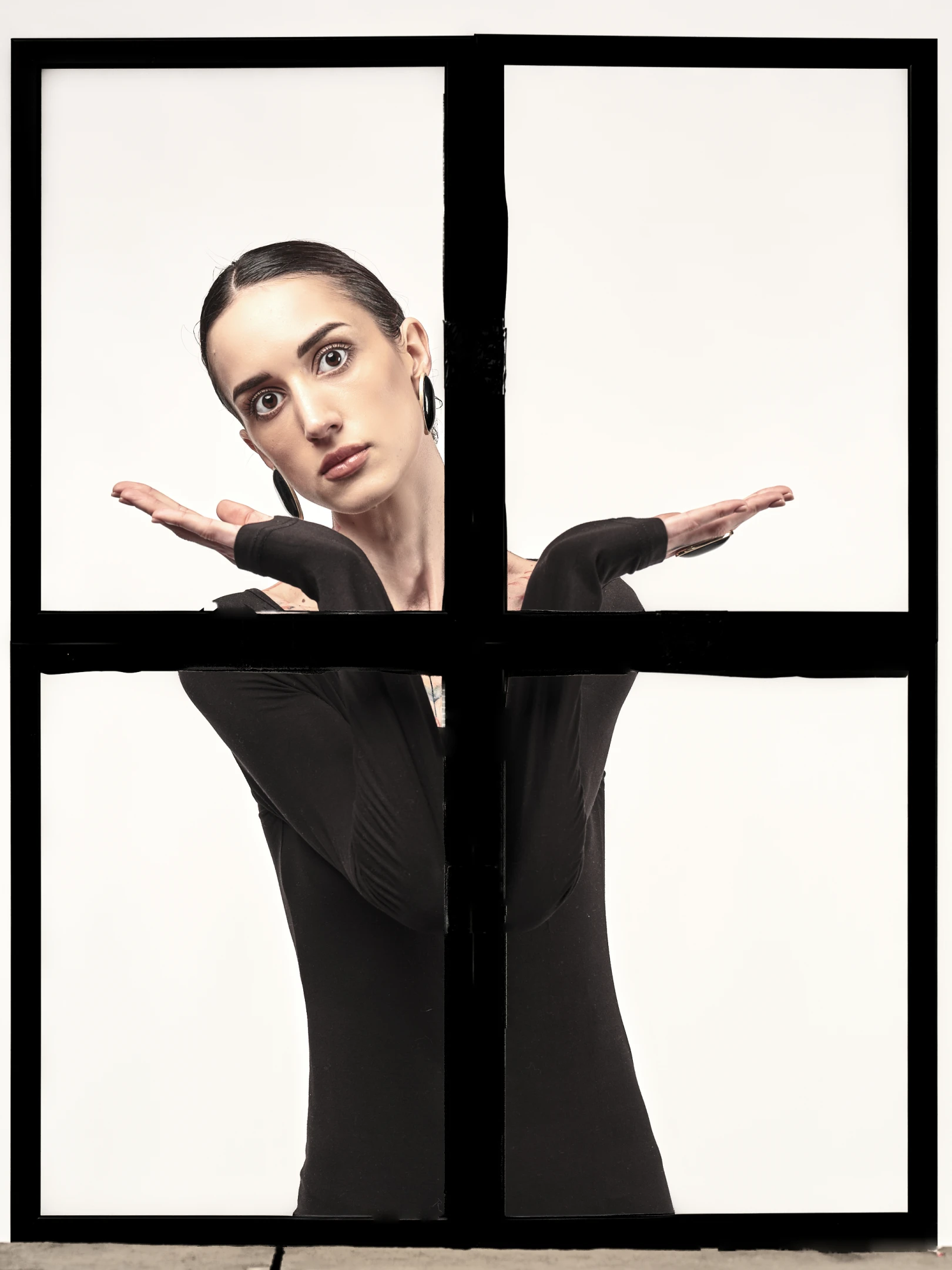 The portrait features a woman with dark, sleekly pulled-back hair, wearing large, dark hoop earrings and a form-fitting black long-sleeve top that accentuates her slender frame. Her expression is direct and intense, with wide, expressive eyes fixed on the viewer, conveying a sense of confidence or defiance. Her arms are extended outward, palms up, as if presenting or offering something, while her body leans slightly into the frame, suggesting a dynamic, almost performative stance. The lighting is high-contrast, with a stark white background that isolates her figure, emphasizing the boldness of her pose and the sharpness of her gaze. The composition is divided into four quadrants by thick ...