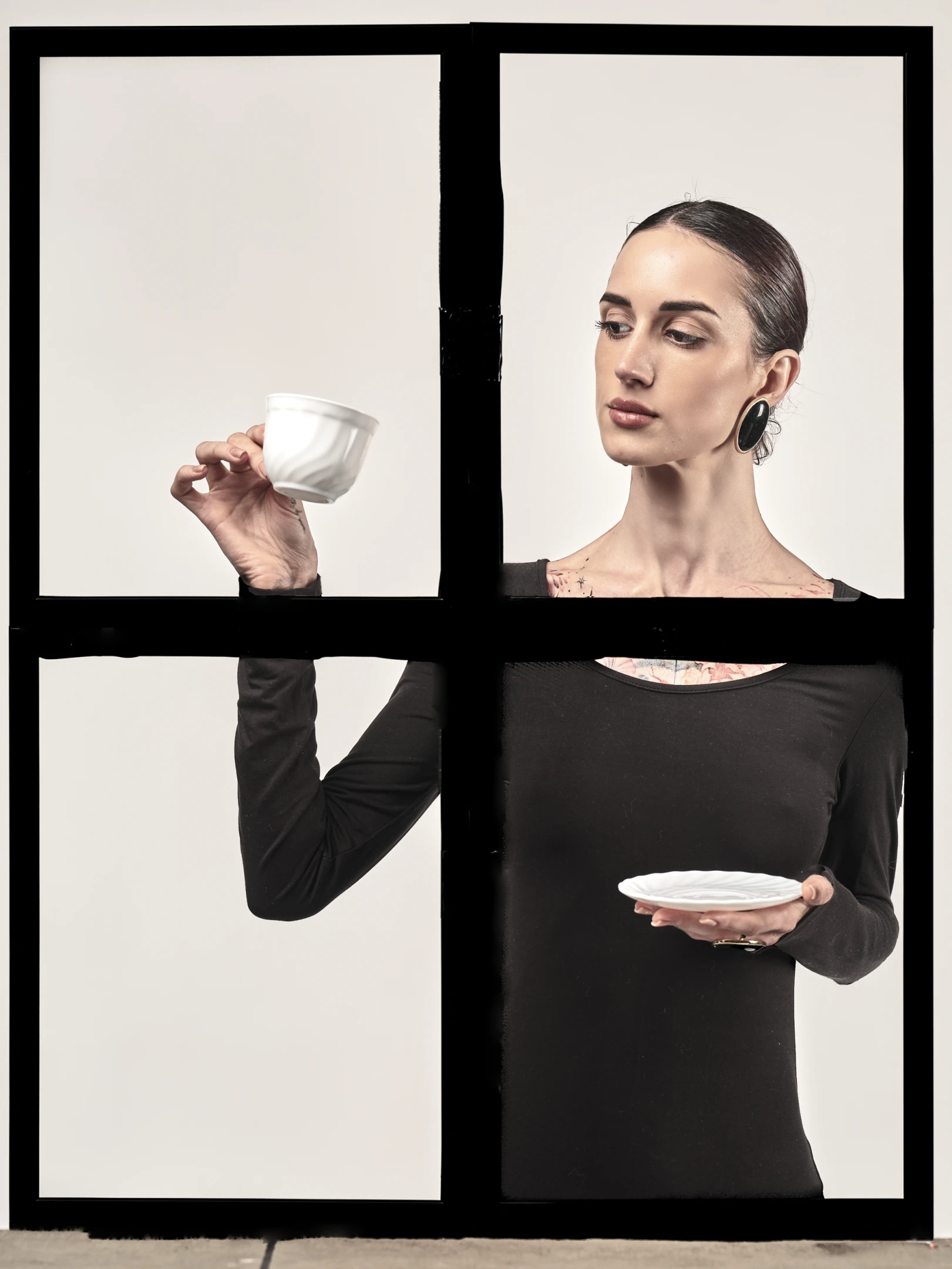 The portrait features a woman with dark, sleek hair pulled back into a neat bun, accentuating her elegant, contemplative expression as she gazes slightly downward with a serene, thoughtful look. Her makeup is minimal yet refined, highlighting her natural features with soft, neutral tones, while large, oval-shaped black earrings add a bold, modern contrast to her otherwise understated look. She wears a fitted black long-sleeve top that reveals a delicate floral tattoo across her collarbone and upper chest, suggesting a blend of sophistication and personal expression. Her pose is composed and deliberate: one hand delicately holds a white ceramic cup suspended midair, while the other cradles...