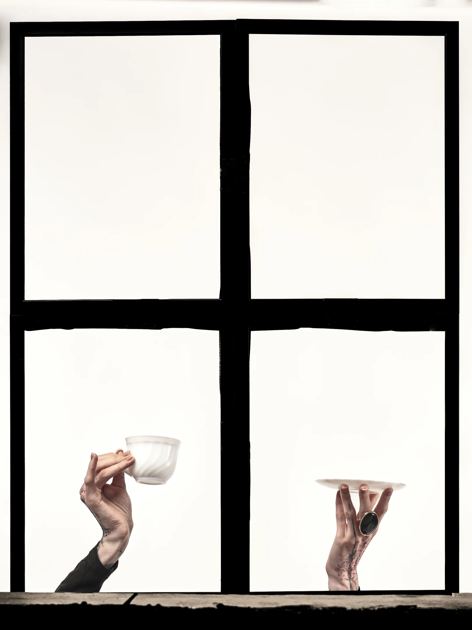 This portrait photograph captures two hands emerging from a stark, minimalist window frame divided into four equal panes, each outlined in bold black. The left hand, adorned with subtle tattoos on the wrist and fingers, delicately holds a small, translucent white cup, while the right hand, wearing a large, dark oval ring and featuring intricate floral tattoos along the forearm, cradles a matching saucer. Both hands are positioned against a uniformly bright, almost ethereal white background, suggesting a studio setting with high-key lighting that casts no shadows, emphasizing purity and clarity. The window’s black frame creates a strong visual contrast, grounding the image in a sense of st...