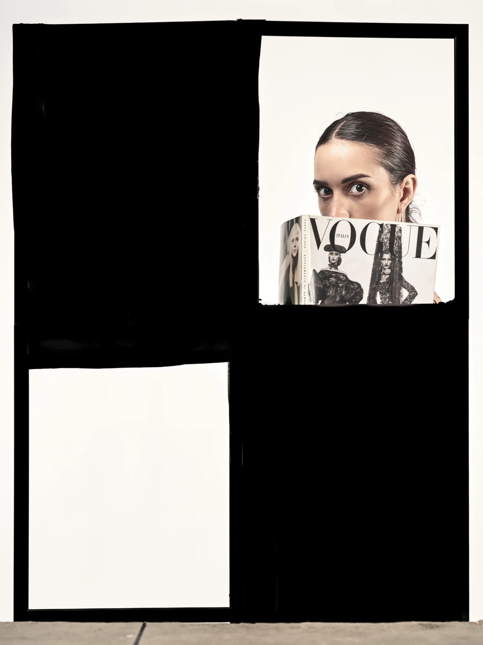 The portrait captures a woman with dark, sleekly pulled-back hair, her wide, expressive eyes gazing directly at the viewer with an air of playful intrigue. She holds a Vogue Italia magazine in front of her face, obscuring her lower features while leaving her piercing gaze and high cheekbones visible, creating a sense of mystery and fashion-forward confidence. Her attire, though partially hidden, suggests a chic, possibly editorial ensemble, with the magazine’s cover featuring classic Vogue styling—black-and-white imagery of models in dramatic poses. The lighting is soft yet focused, highlighting her facial features against a stark, minimalist background that emphasizes the subject’s prese...