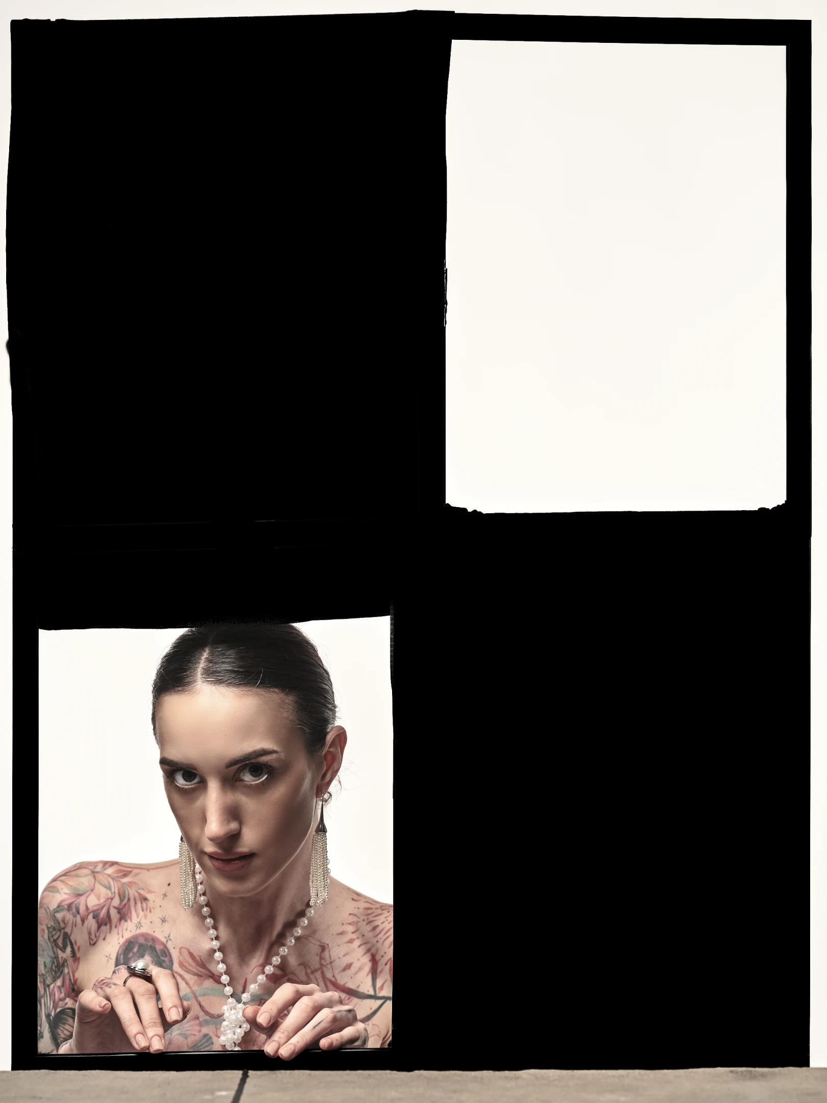 The portrait captures a woman with dark, neatly pulled-back hair, gazing directly at the viewer with an intense, almost confrontational stare. Her skin is adorned with intricate, colorful tattoos covering her shoulders, chest, and arms, featuring floral and abstract patterns with red and black tones. She wears a delicate pearl necklace and long, dangling pearl earrings, adding a touch of elegance that contrasts with her bold tattoos. Her hands, adorned with rings, rest gently on the edge of the frame, fingers slightly curled as if holding or presenting something unseen. The lighting is soft yet focused, highlighting her facial features and the textures of her tattoos and jewelry, while ca...