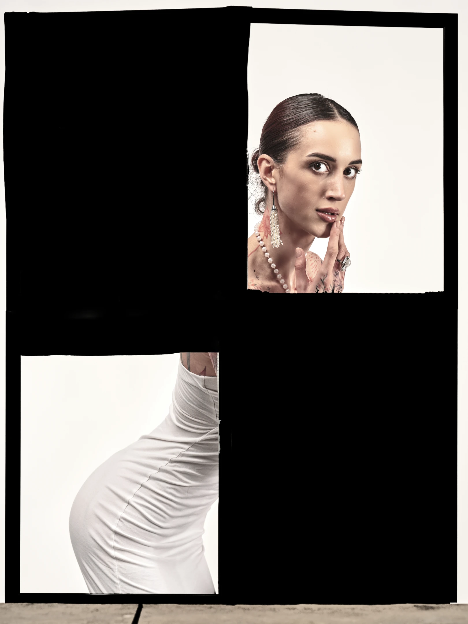 The portrait features a woman with dark, sleekly pulled-back hair, gazing directly at the viewer with an intense, captivating expression. Her hand gently touches her cheek, fingers adorned with rings and tattoos, while a delicate pearl necklace and long, dangling earrings accentuate her neck and shoulders. She wears a form-fitting white dress that hugs her curves, emphasizing the smooth texture and ruched lines of the fabric. The lighting is soft yet dramatic, highlighting her facial features and the contours of her dress, creating a mood of quiet confidence and allure. The background is stark white, isolating her figure and enhancing the contrast with the deep black borders that frame th...