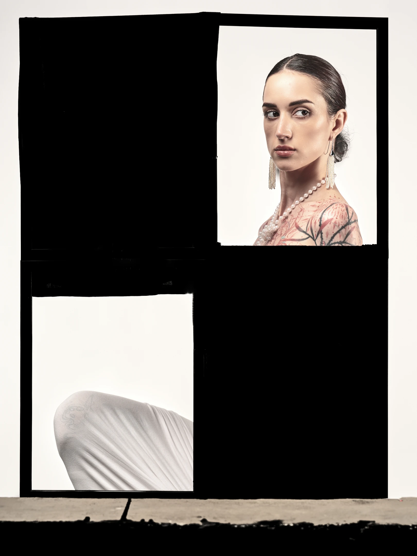 The portrait features a woman with dark, sleek hair pulled back into a neat bun, revealing a poised, contemplative expression as she gazes slightly off-camera with a calm, introspective look. Her skin is fair, and she wears a delicate strand of pearls around her neck, complemented by long, dangling earrings that shimmer with fine, cascading beads. Her shoulders and upper arms are adorned with intricate tattoos—featuring floral and leaf-like patterns in soft pink and black tones—adding a layer of personal artistry to her appearance. She is dressed in a flowing, white garment that drapes elegantly, suggesting a sense of grace and movement, though the image captures only a portion of her for...