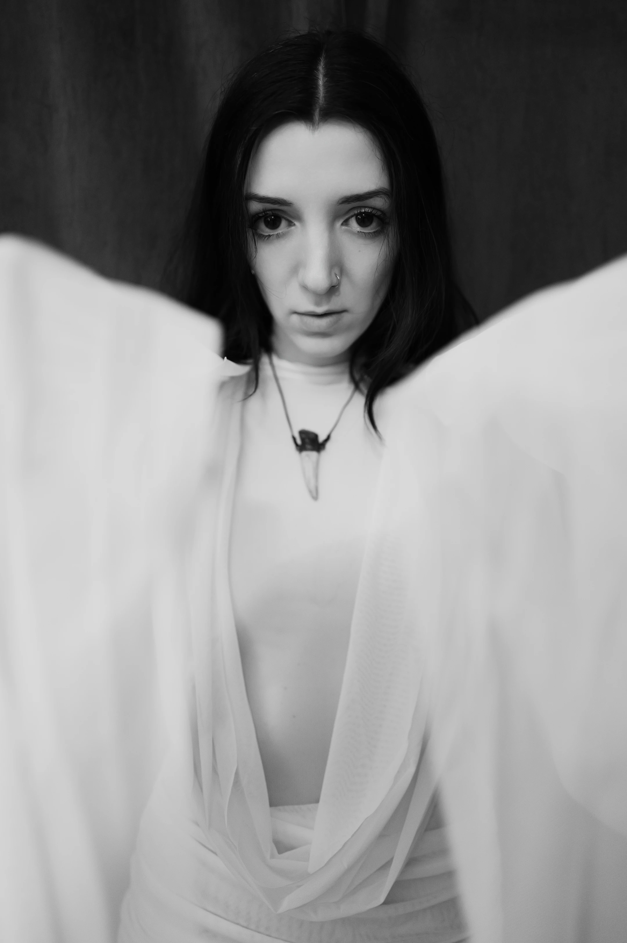 This is a striking black-and-white portrait of a young woman with long, dark hair parted down the middle, gazing directly into the camera with a calm, intense expression. Her face is framed by a flowing, translucent white fabric that drapes elegantly around her shoulders and torso, creating a sense of ethereal grace and movement. She wears a simple white top beneath the fabric, accessorized with a delicate necklace featuring a sharp, pointed pendant. Her expression is neutral yet compelling, with wide, dark eyes and a subtle nose piercing adding to her enigmatic presence.