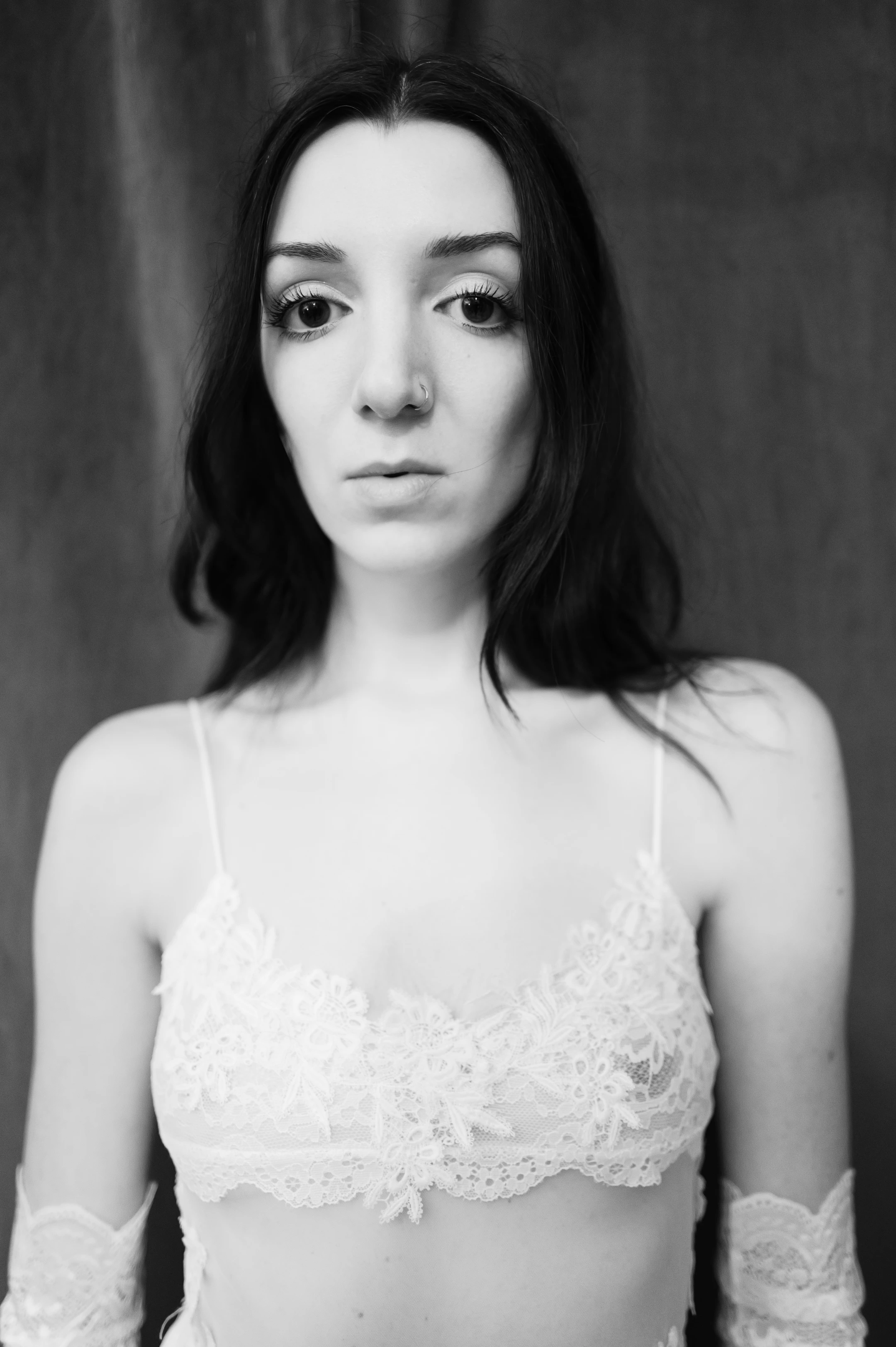 A black and white portrait of a woman with shoulder-length dark hair and a nose ring, gazing directly at the camera with a neutral expression. She wears a delicate white lace bralette with thin straps and matching lace arm warmers. The lighting is soft and frontal, creating gentle highlights on her skin and the intricate lace patterns, while the dark, blurred background isolates her figure. The overall mood is intimate and artistic, with a timeless, contemplative quality.