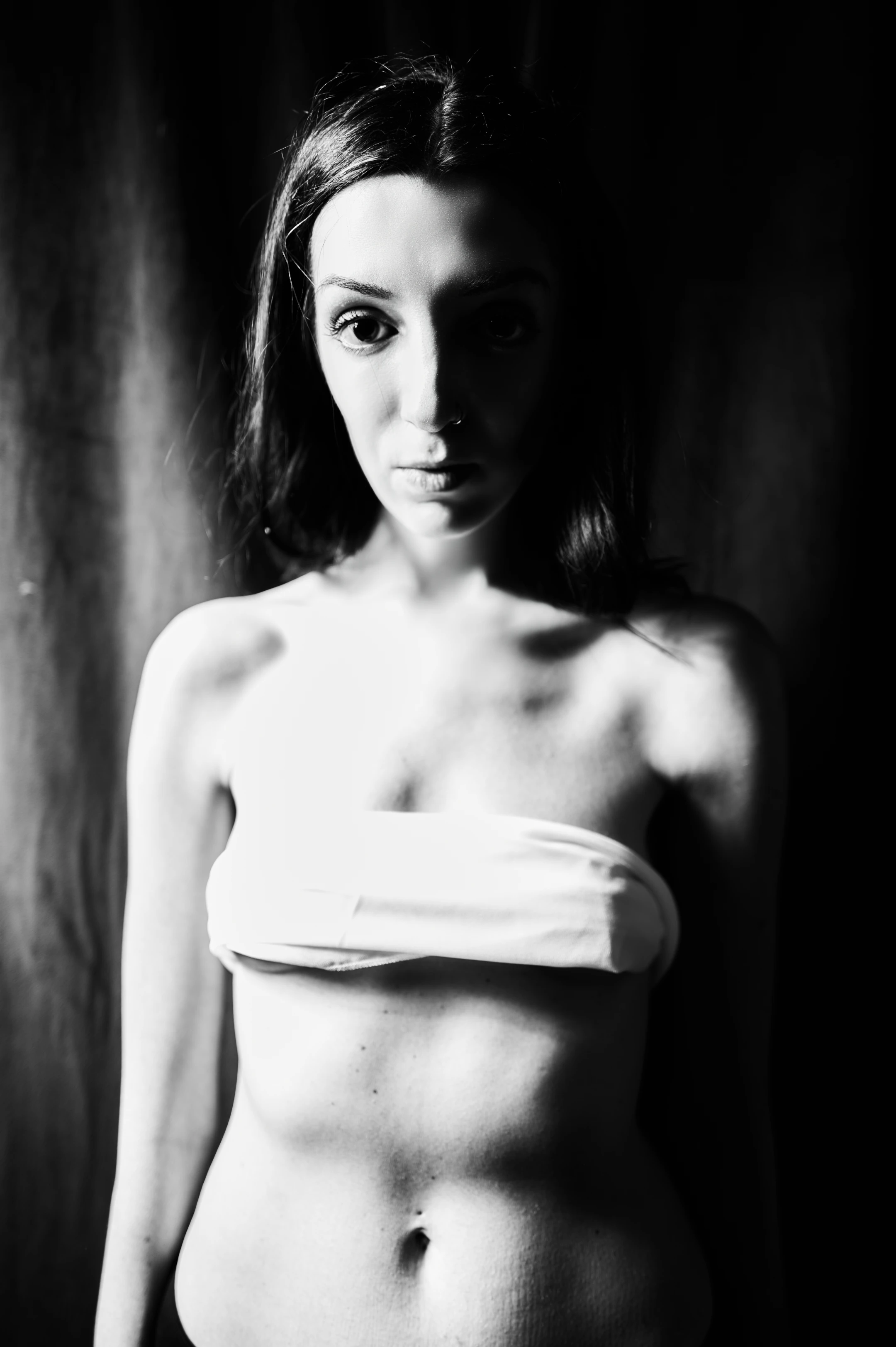 This is a high-contrast black-and-white portrait of a woman standing center-frame against a dark, textured backdrop that appears to be a curtain or draped fabric. Her long, dark hair frames her face, which is illuminated from the front, casting deep shadows behind her and accentuating her piercing gaze and neutral, intense expression. She is bare-chested, wearing a simple white bandeau that wraps snugly around her bust, leaving her abdomen and lower torso exposed. Her arms are slightly away from her body, and she stares directly into the camera with a calm, almost confrontational intensity.